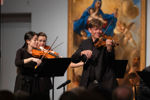 A violinist in front of a religious inspired painting, surrounded by other orchestra players.