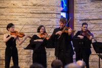 A violinist is performing, surrounded by other orchestra players against a tan wall.
