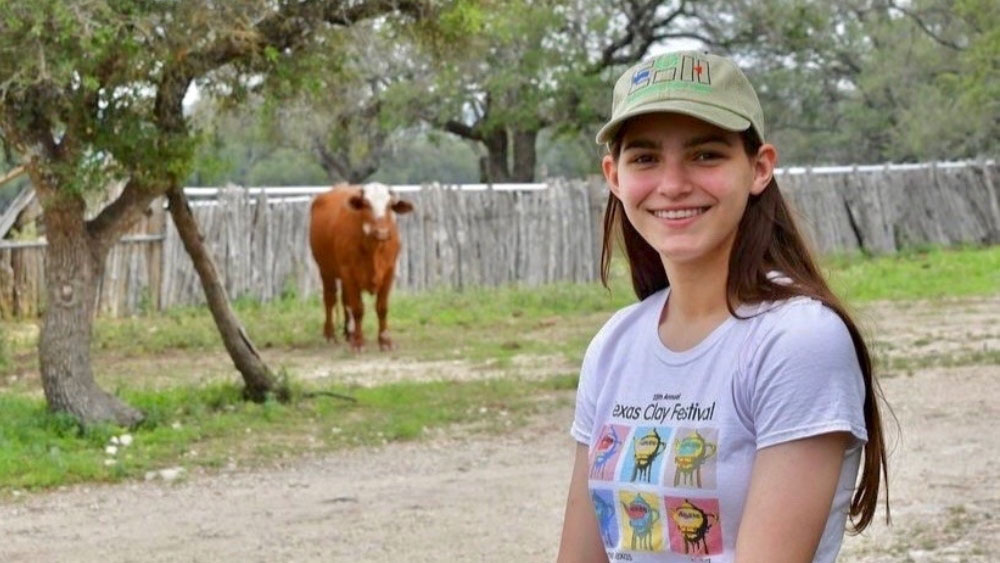 Miranda Duggan An individual wearing a cap. In the background, a solitary cow stands in a field surrounded by trees and a fence line.