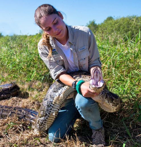 Jaimie is kneeling on the ground while holding a python behind its head.