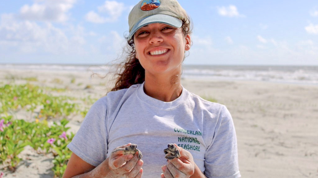 Isabel is standing on a beach holding two turtle hatchlings, one in each hand. She is smiling at the camera.