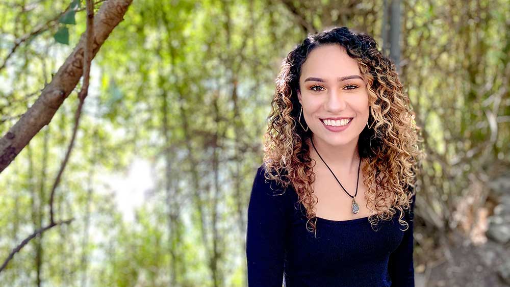 Tayler is standing in front of a vibrant green forest. She is smiling at the camera.