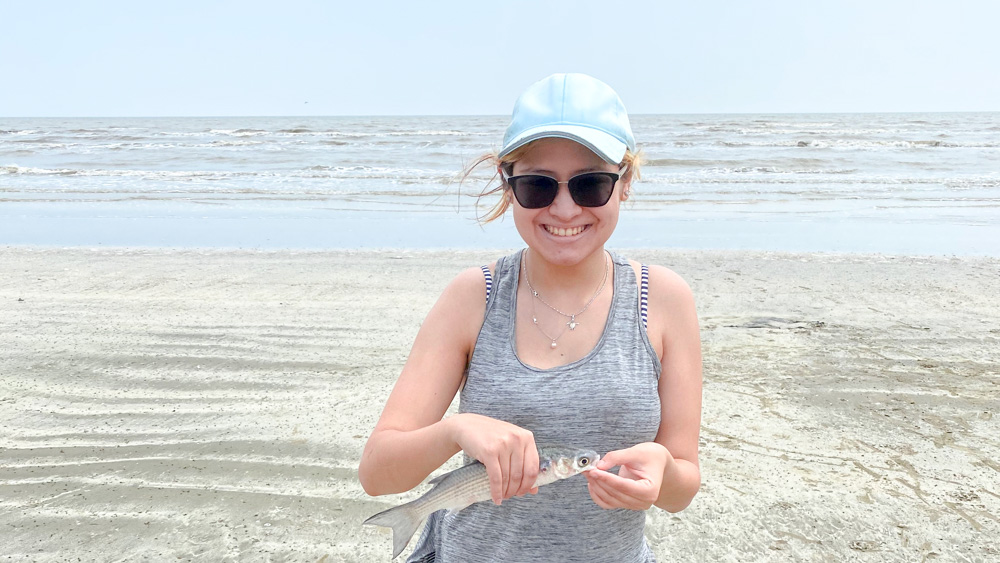 Victoria is on the beach holding a fish with both hands.