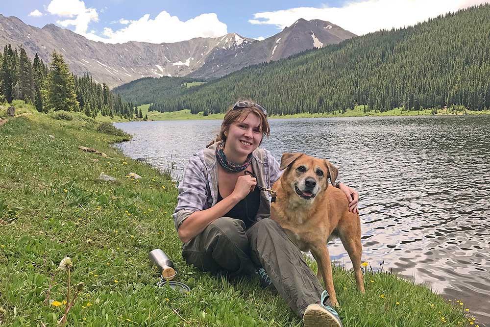 Sarah is sitting on a grassy bank of a lake with her dog with an evergreen forest and mountains in the background.