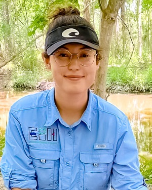 Aurora is wearing a black sun visor and smiling at the camera. A stream and trees are in the background.