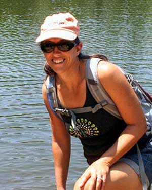 Kristi is standing in front of a large body of water and smiling to the camera. She is wearing a cap, sunglasses, and field clothes.