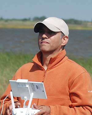Marc looks into the distnce. He is wearing a cap and an orange sweatshirt. He is holding a drone controller. Marsh grasses and a body of water are in the background.