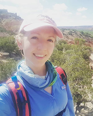 Kaylei is wearing a pink cap and field clothes. She is smiling at the camera. A rocky hillside and trees are in the background.