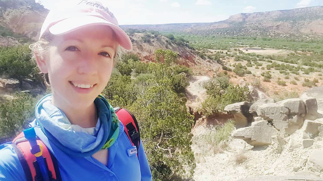 Kaylei stands in front of a scenic view of rugged terrain with greenery and distant hills.