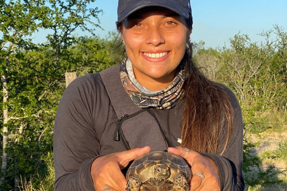 Brandi is in the field. She is holding a tortoise and they are both smiling at the camera.