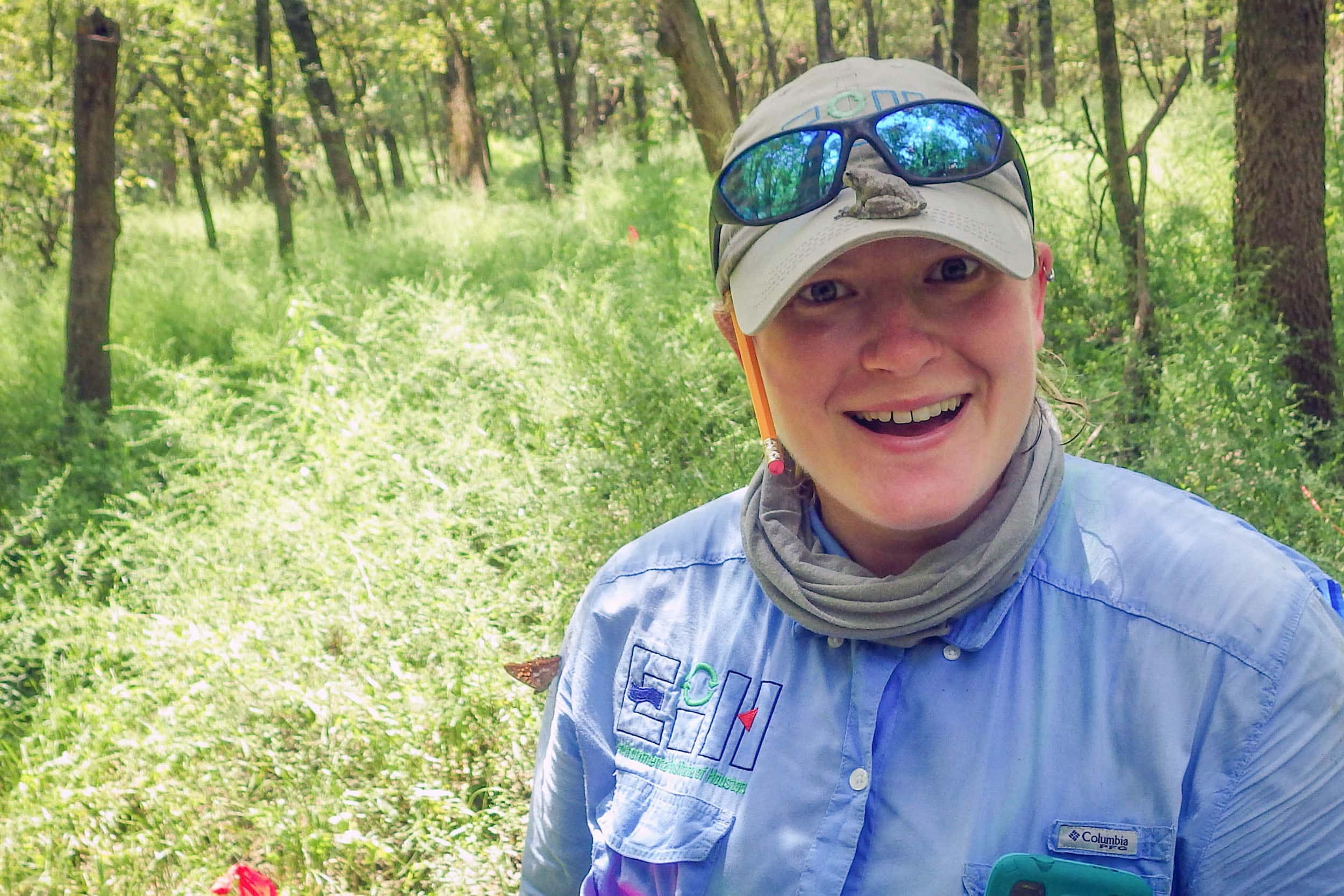 Mandi is outside and is smiling into the camera. She is amused by the gray tree frog sitting on the bill of her khaki-colored cap. The background is filled with bright green plants and dark brown tree trunks.