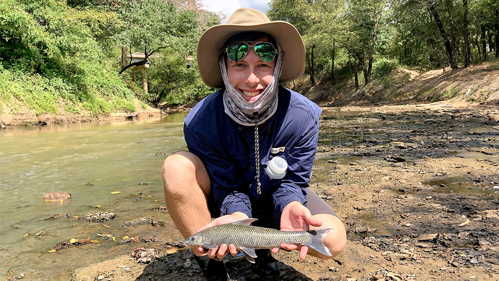 Noah is crouched in a stream bed, holding a fish and smiling at the camera.