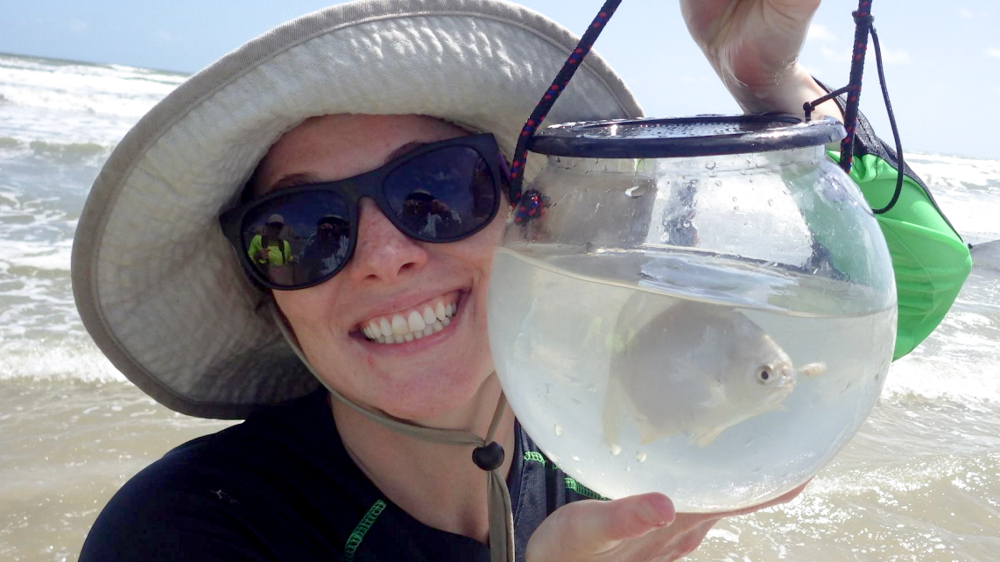 Emily is holding an observation tank with a silver fish up to her face. She is smiling at the camera.