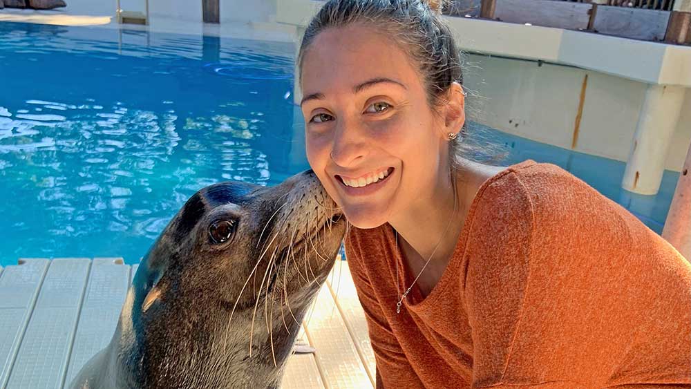 Kelly is smiling at the camera while a sea lion is giving her a kiss on the cheek.