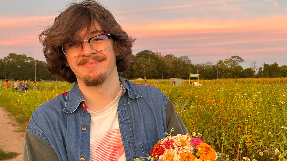 Luke happily stands in a field of flowers, holding a colorful bouquet. He smiles at the camera against a stunning blue and pink sky.