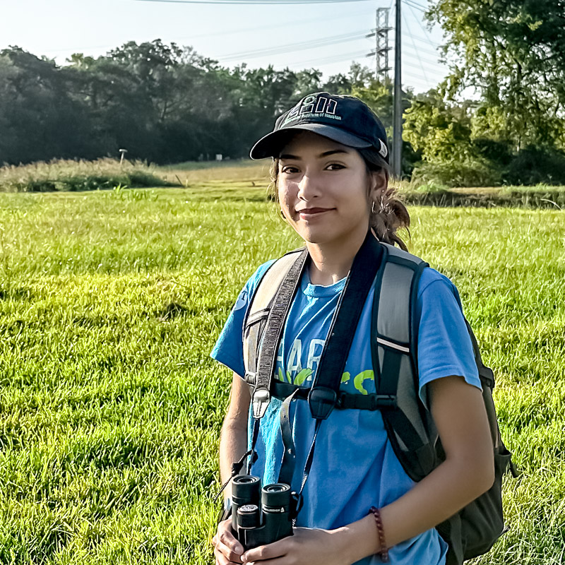 Keilah stands in a sunny field with birdwatching gear.