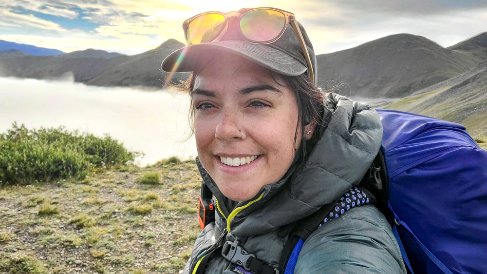 A person in outdoor gear stands in front of a scenic backdrop of mist-covered mountains.