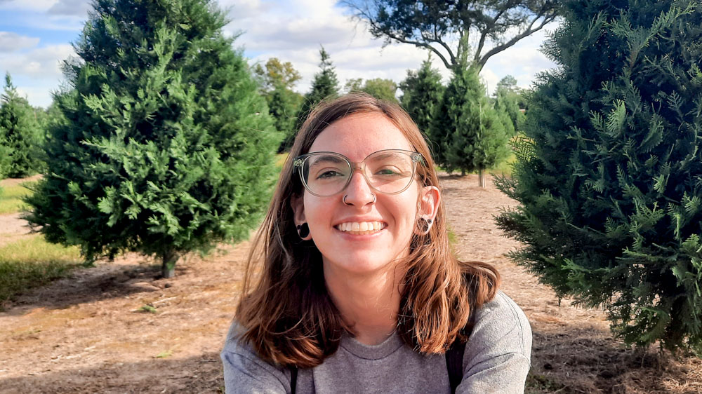 Jasmine is smilling at the camera and is surrounded by pine trees.