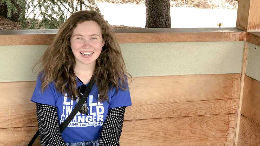 Tori is sitting on a natural wood bench and is smiling at the camera.