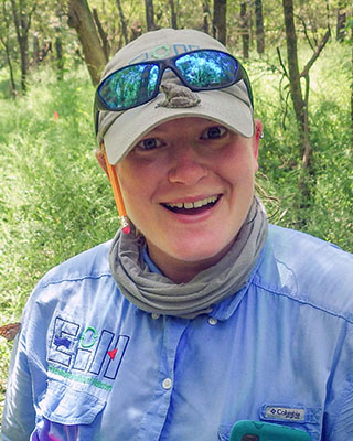 Mandi is outside and is smiling into the camera. She is amused by the gray tree frog sitting on the bill of her khaki-colored cap.