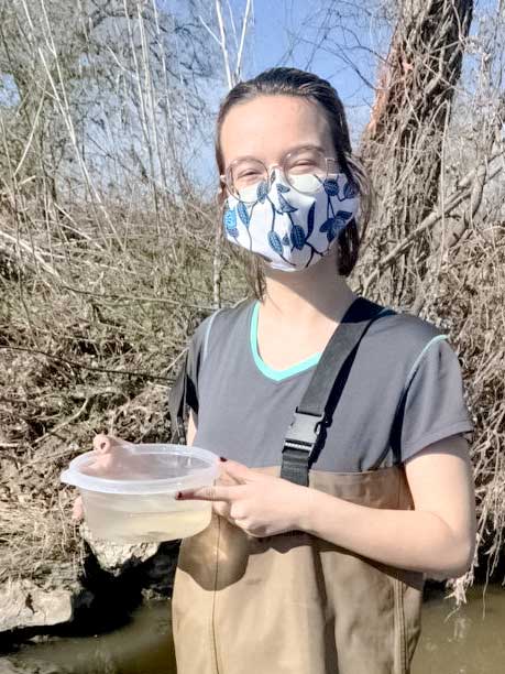 Catherine is masked up and holding a container filled with water and a few small fish. In the background is a stream bank covered in bare stems.