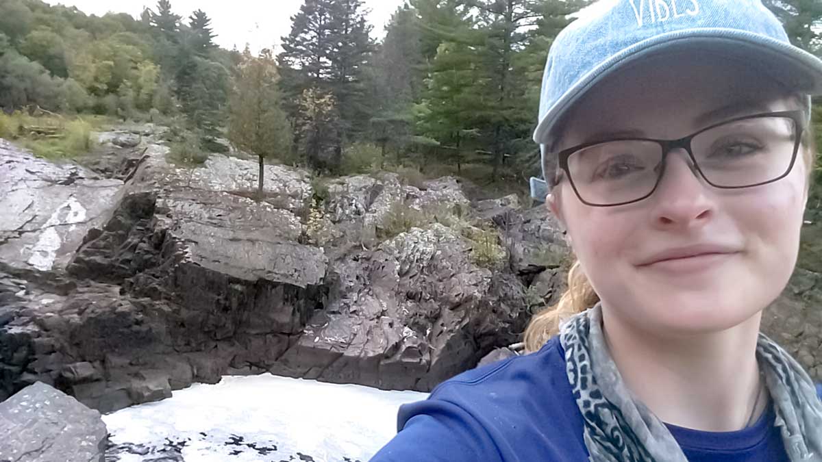 Jenna is smiling into the camera. Behind her is a rocky slope with snow at the bottom and green trees above.