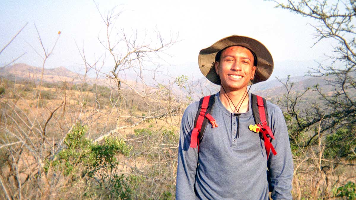 Garret is in full field gear standing among some brush. He is smiling into the camera.