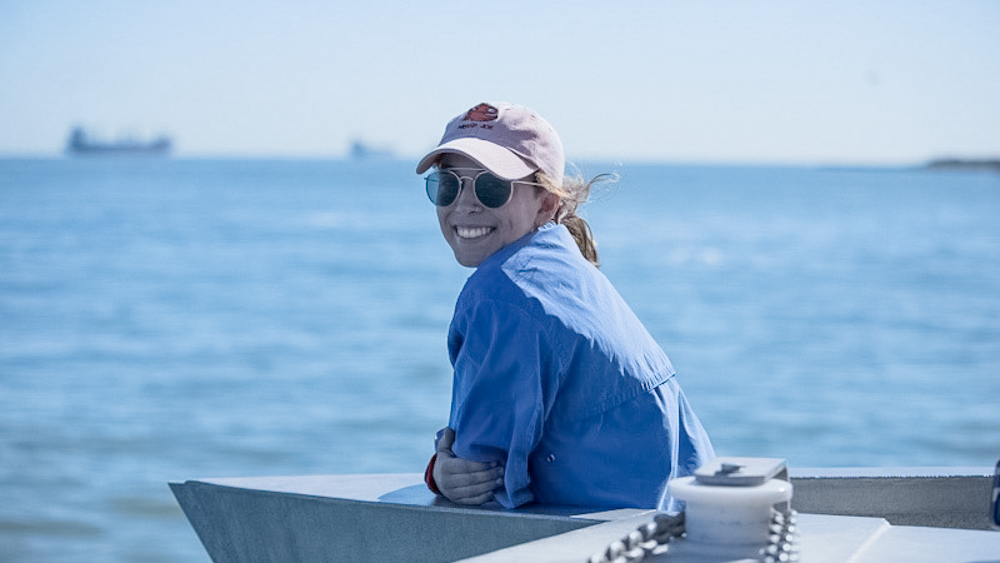 Taryn is on a ship and smiling at the camera with the ocean and distant ships in the background.