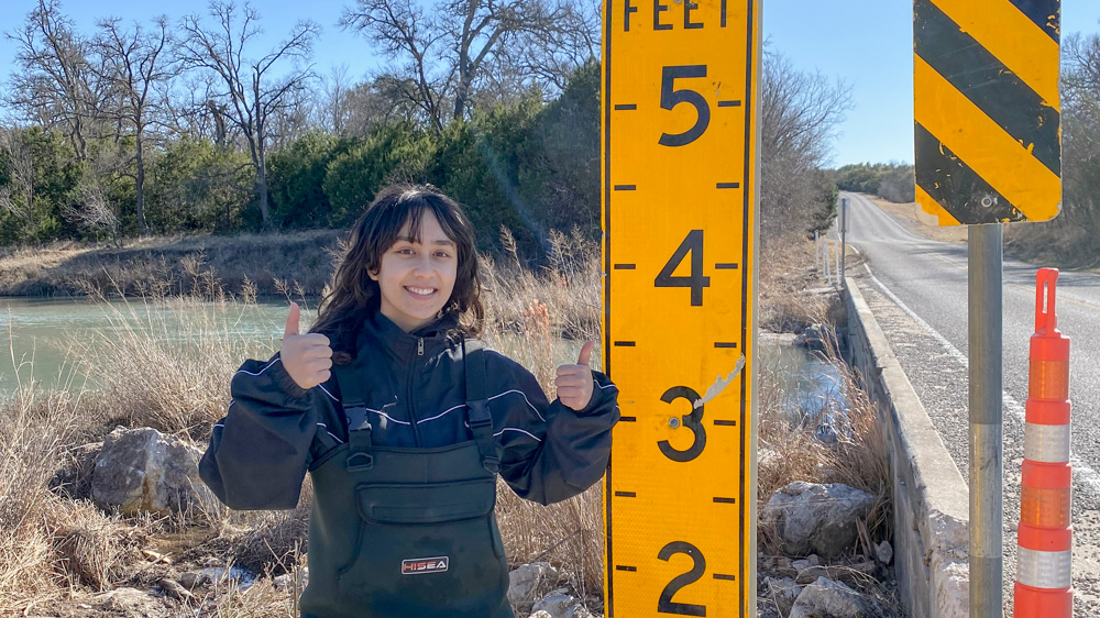 Adriana is giving two thumbs up from where she stands next to a flood gauge.