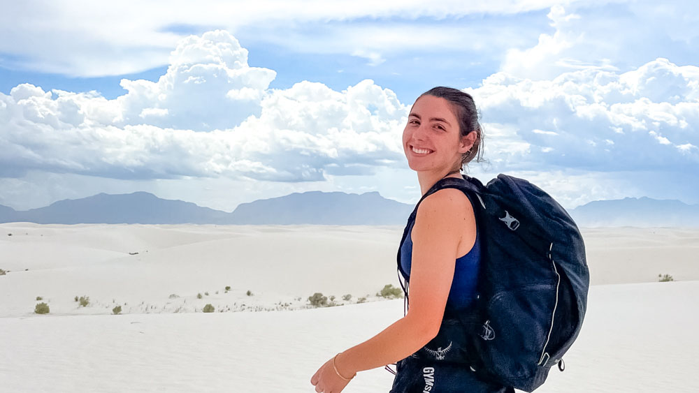 Ashlyn Sak is hiking on sand dunes. There are fluffy white clouds in the sky and mountains in the distance. She is carrying a packback and smiling at the camera over her shoulder. Ashlyn Sak