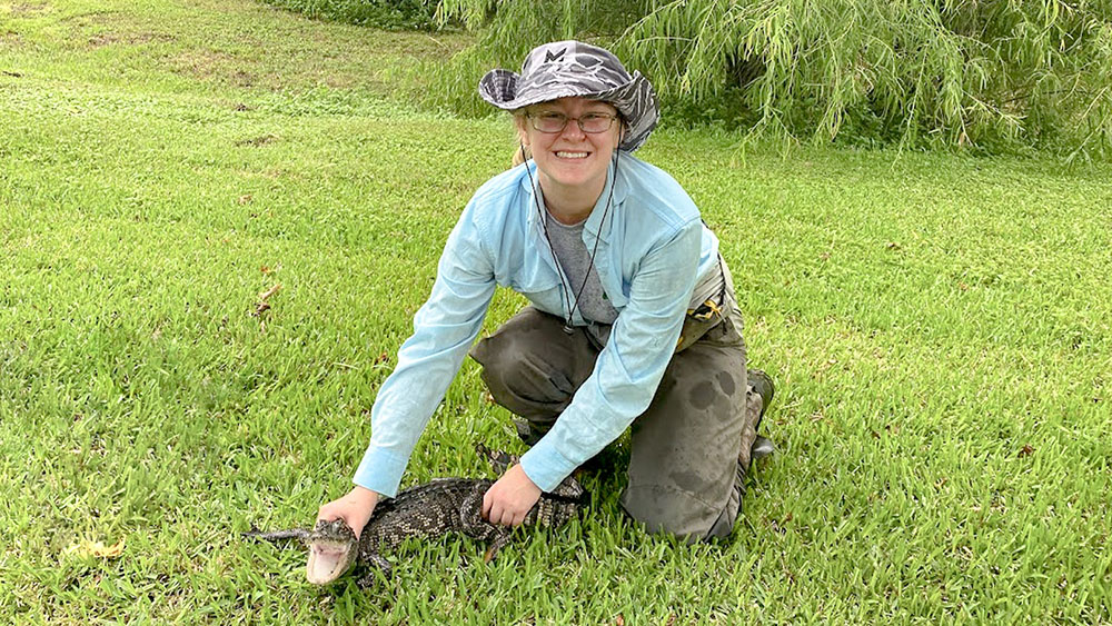 Taylor is kneeling on one knee while holding down a small alligator.