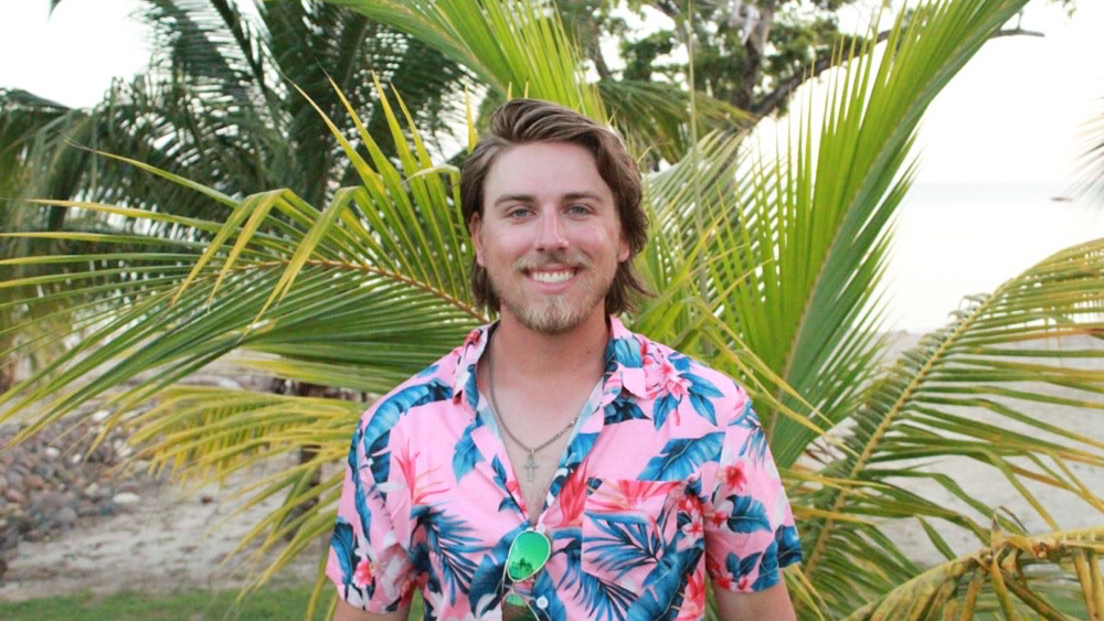 Joshua is standing in front of a palm tree on beach. He is wearing a colorful Hawaiian shirt and smiling at the camera.