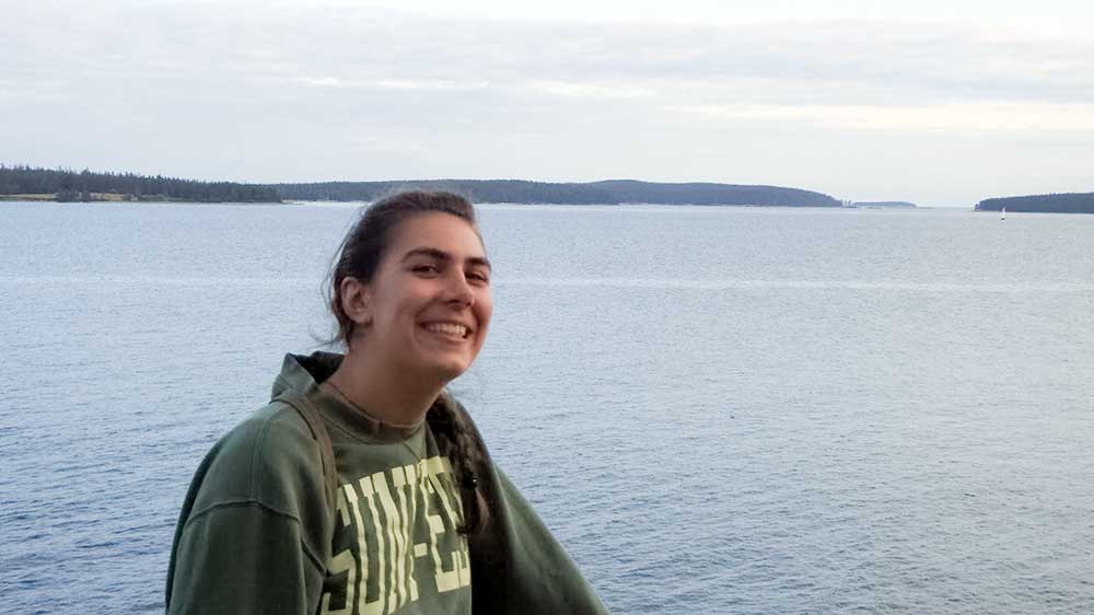 Haley is smiling into the camera. Behind her is a large body of water with tree-covered hills in the horizon.