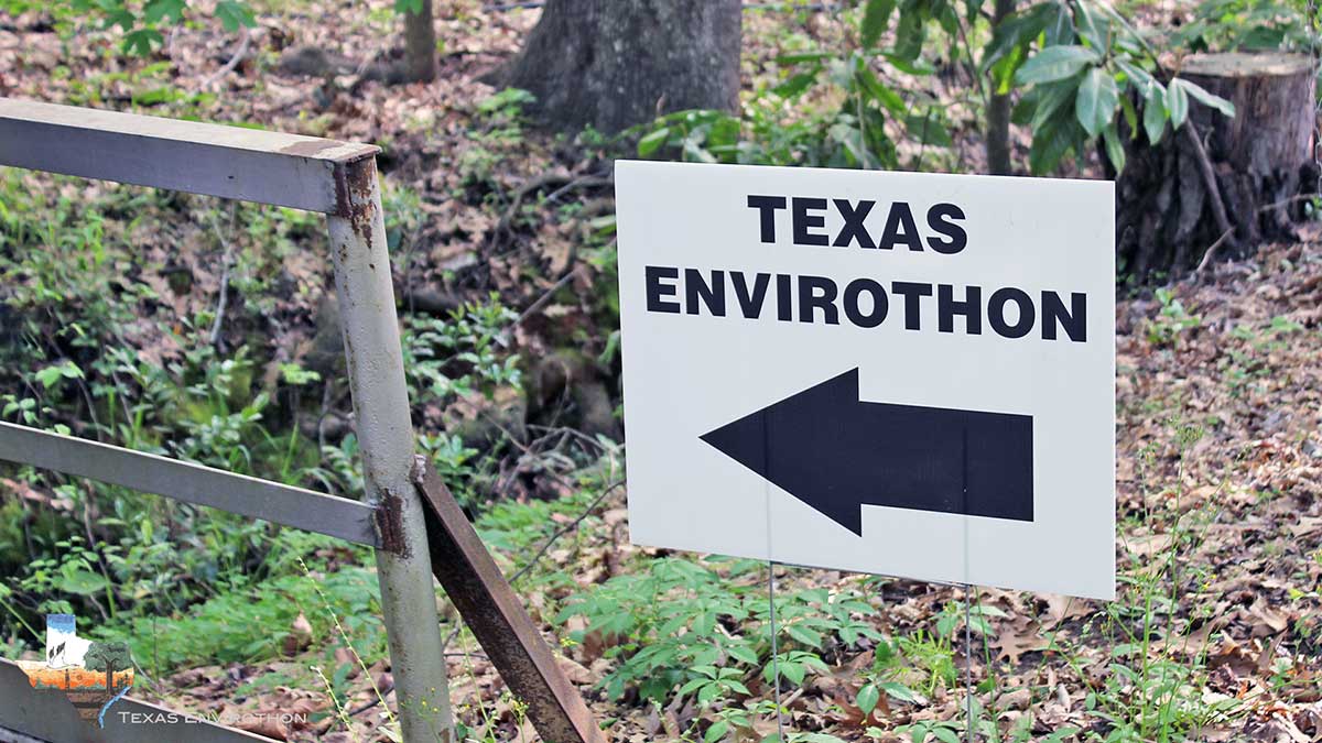 White sign in the woods with black left-pointing arrow showing the way to the Texas Envirothon.