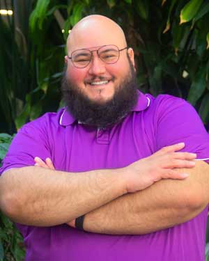 Portrait photo of Joshua Quinn wearing a purple photo and purple rimmed glasses.